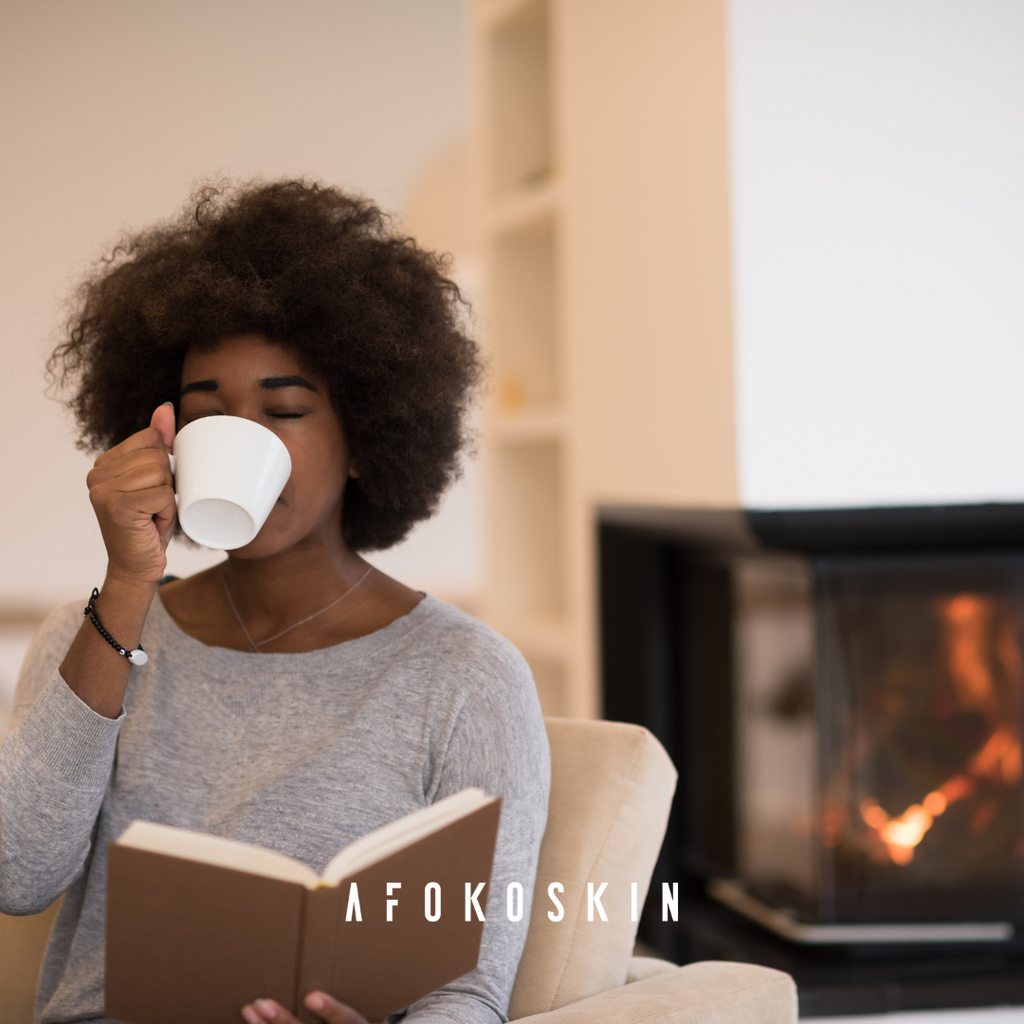 This photo shows a black woman with natural hair sipping tea and reading a book by the fire.